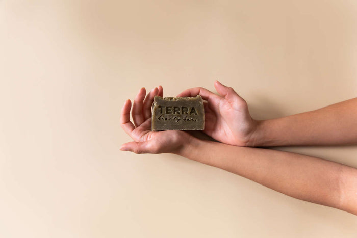Two hands hold a Clean Green Tea Tree Facial Beauty Bar by Terra Beauty Bars against a beige backdrop. Embossed with TERRA and infused with Green Sea clay, the natural, handcrafted soap is ideal for skincare enthusiasts who value purity.