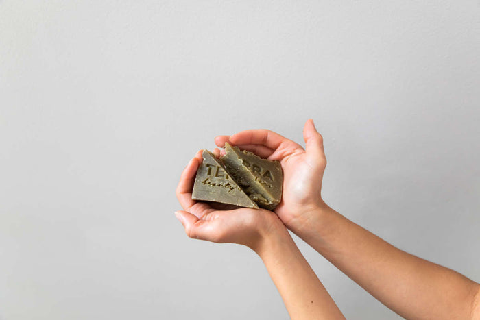 Gently held in two hands, a cracked Terra Beauty Bars Clean Green Tea Tree Facial Beauty Bar features engraved text. Its matcha tea-infused earthy tones contrast with the light beige background, creating a minimalist and soothing composition.