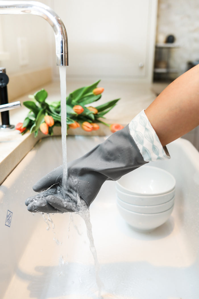 In a picturesque scene, someone wearing durable Grove Co. Cleaning Gloves rinses their hand under running water in the kitchen sink. Nearby, three stacked white bowls and a bouquet of orange tulips add a touch of elegance to this sustainable cleaning moment.