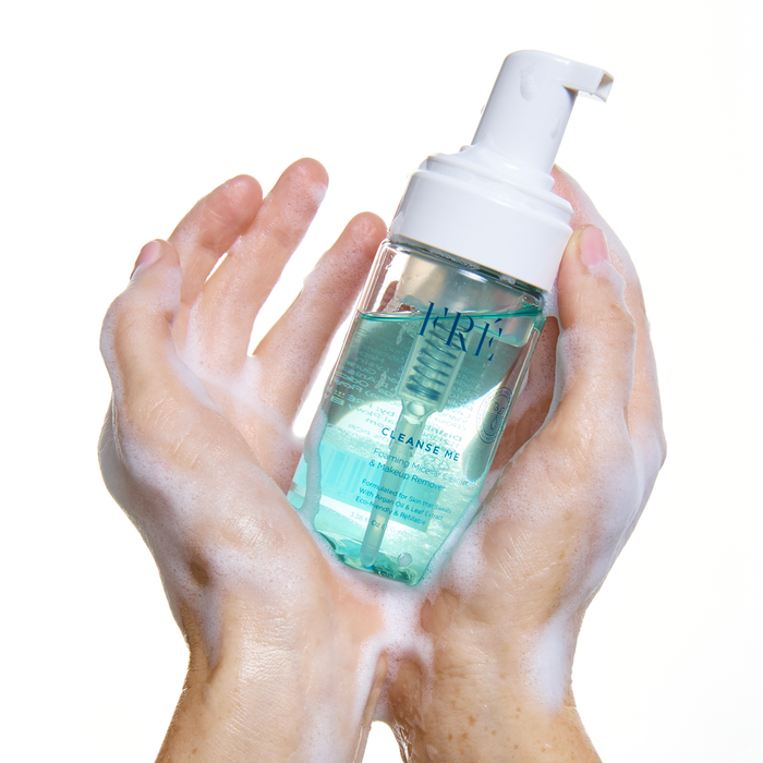 A pair of soapy hands holds CLEANSE ME Foaming Micellar Water by FRÉ Skincare, a clear bottle with a white pump dispenser and blue cleanser inside, against a white background.