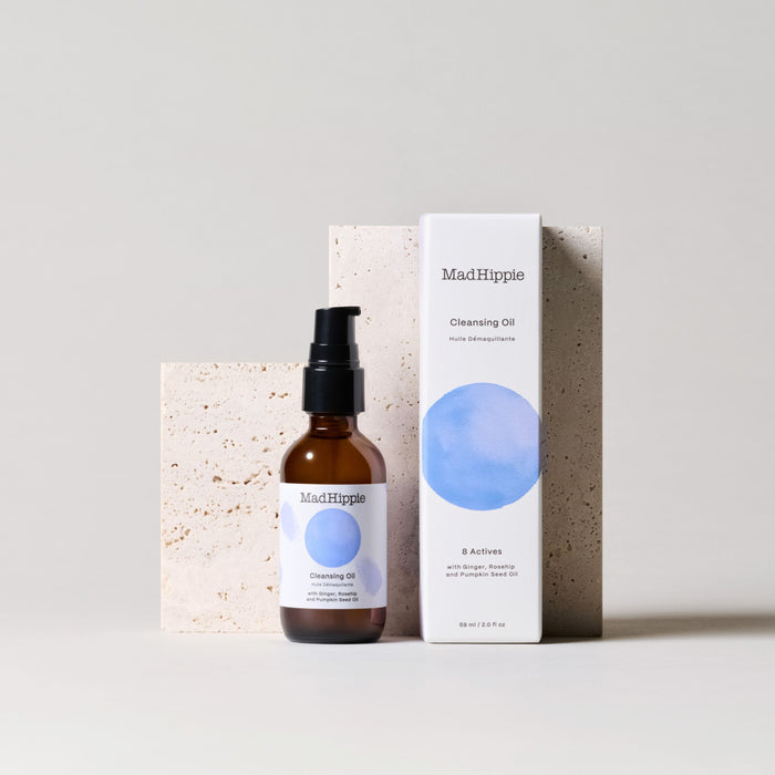 Mad Hippies Cleansing Oil with organic pumpkin seed oil is shown in an amber bottle with a black pump and a box featuring a blue circle design, set against a beige background with textured stone, highlighting its place in natural skin care.