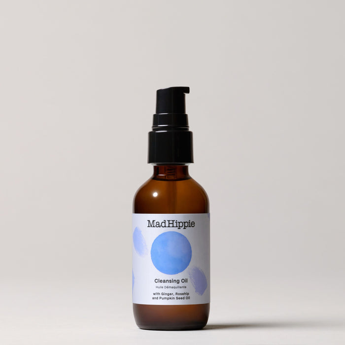 A dark amber pump bottle, Cleansing Oil by Mad Hippie, sits against a light background. The label features blue circular designs and boasts organic ingredients such as ginger, rosehip, and pumpkin seed oil—ideal for natural skincare enthusiasts.