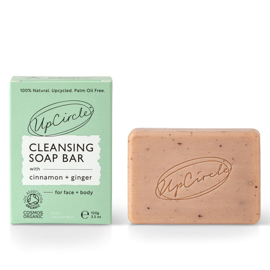 The UpCircle light brown Cleansing Soap Bar with Cinnamon + Ginger features visible specks and offers gentle exfoliation for radiant skin. It sits beside its light green packaging, proudly displaying the UpCircle brand name.