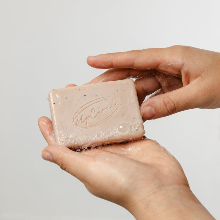 Two hands hold a lathered Cleansing Soap Bar with Cinnamon + Ginger from UpCircle. The light pink bar, infused with pink clay and flecked, displays an engraved logo, ideal for gentle exfoliation and promising a radiant complexion against a plain white background.