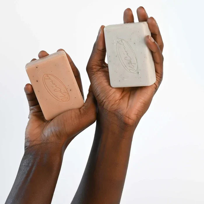Two hands present rectangular UpCircle soap bars against a plain background: one is the Cleansing Soap Bar with Cinnamon + Ginger, and the other is infused with pink clay for gentle exfoliation. Both feature an embossed design, highlighting these soaps as a step towards a radiant complexion.