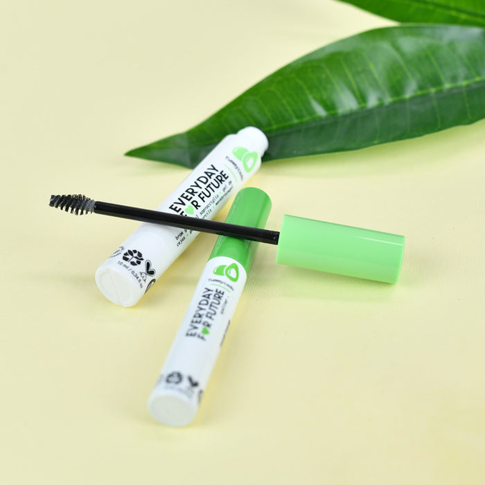 Two tubes of Clear Avocado Brow Gel by Everyday For Future rest on a light yellow surface. One tube is open, displaying a black brush. These vegan, cruelty-free delights use organic ingredients. A lush green leaf in the background enhances their natural appeal with green caps on display.