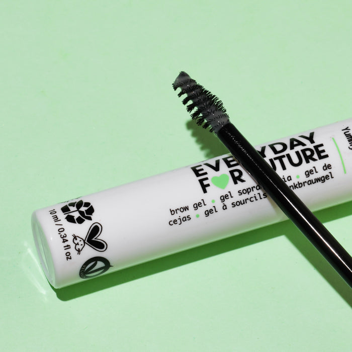 A sleek white tube of Everyday For Futures Clear Avocado Brow Gel with Castor Oil sits on a light green backdrop. A black spoolie brush infused with organic avocado oil is placed diagonally atop it, highlighting its vegan and cruelty-free icons.