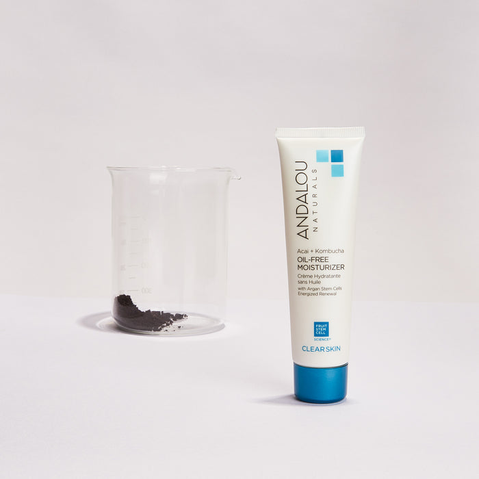 A tube of Andalou Naturals Clear Skin Acai + Kombucha Oil-Free Moisturizer stands upright against a light backdrop. A nearby beaker holds some black powder, emphasizing the fruit stem cell complex for effective nourishment and clear skin benefits.