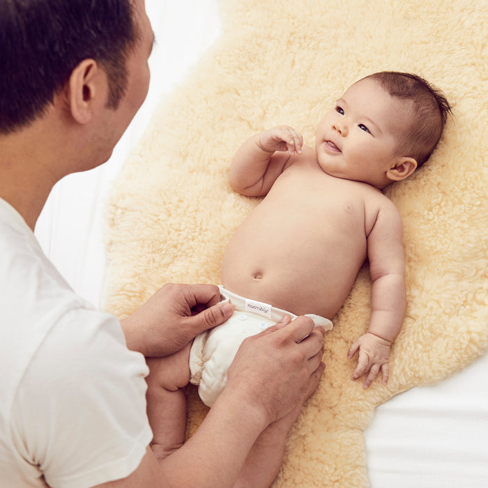 An adult wearing a white shirt gently changes a smiling baby on a soft cream blanket. The content baby lies on their back in an Esembly Baby Size 1 Cloth Diaper Inner, made from 100% OEKO-TEX Certified unbleached organic cotton, designed to prevent blowouts.