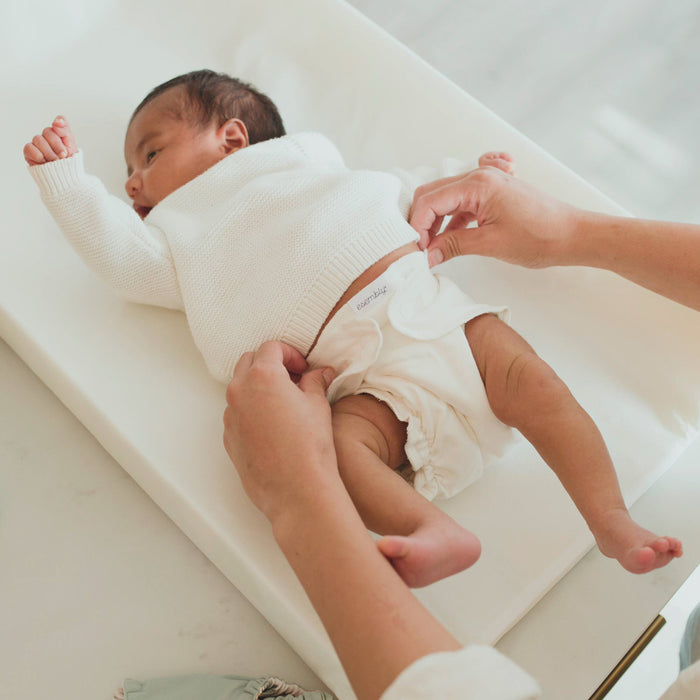 A baby in a white sweater lies on a changing table as someone adjusts an Esembly Baby Cloth Diaper Inner, Size 1, made from 100% OEKO-TEX Certified Unbleached Organic Cotton. The babys left arm is slightly raised amidst a bright setting with a neutral background, enhancing the organic cotton softness.