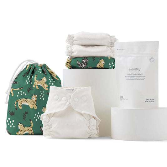 Esembly Babys Cloth Diaper Try-It Kit, Size 1 includes a cream cloth diaper with snaps, a cheetah-patterned green organic cotton drawstring bag, folded white cloths, and washing powder, elegantly displayed on cylindrical white stands.