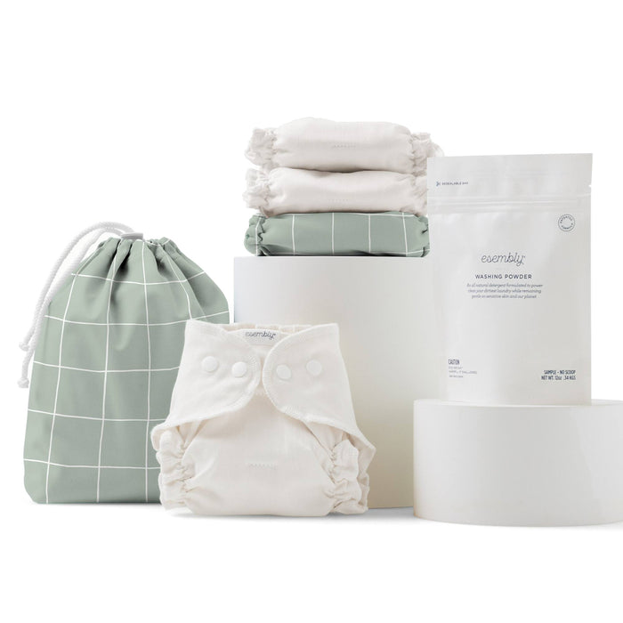The Esembly Baby Cloth Diaper Try-It Kit, Size 1, features a stylish ensemble of eco-friendly items: three organic cotton cloth diapers, one fastener-equipped diaper, a white packet of washing powder, and a green drawstring bag with a white grid pattern arranged elegantly on cylindrical stands.