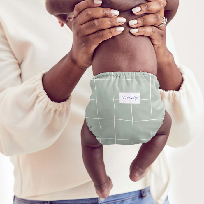 An adult holds a baby wearing Esembly Babys eco-friendly Cloth Diaper Try-It Kit, Size 1, featuring a green and white checkered design with text. Made from organic cotton, it pairs perfectly with the adults cream sweater as they share a cherished moment.