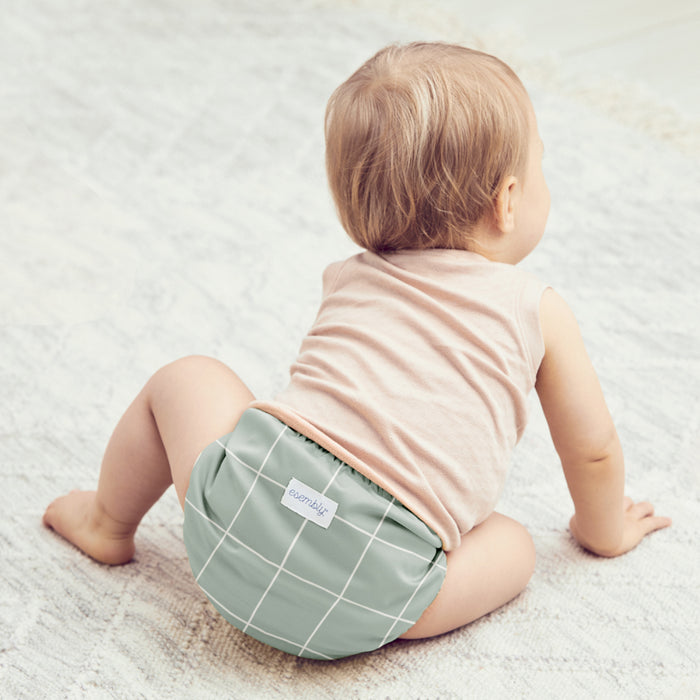 An infant in an eco-friendly green, grid-patterned Esembly Baby Cloth Diaper Try-It Kit, Size 1, sits on a soft beige carpet. Facing away, they gaze rightward, showcasing charming sustainable style in a sleeveless shirt.