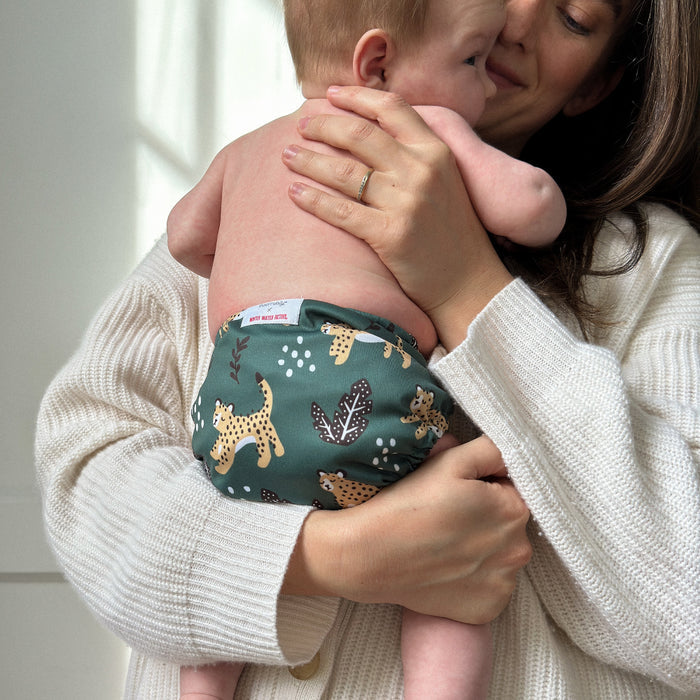 A woman in a white sweater holds a baby wearing Esembly Babys Cloth Diaper Try-It Kit, Size 1, crafted from organic cotton with cheetah and leaf prints. The baby rests gently against her shoulder in an eco-conscious, softly lit room.