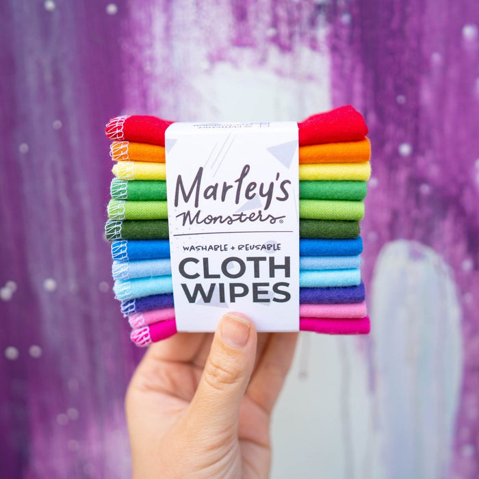 A hand holds a pack of Marleys Monsters Cloth Wipes against a purple and white backdrop. The neatly stacked, vibrantly rainbow-colored wipes are made from soft cotton flannel. The label proudly states washable + reusable cloth wipes, highlighting their handmade quality.