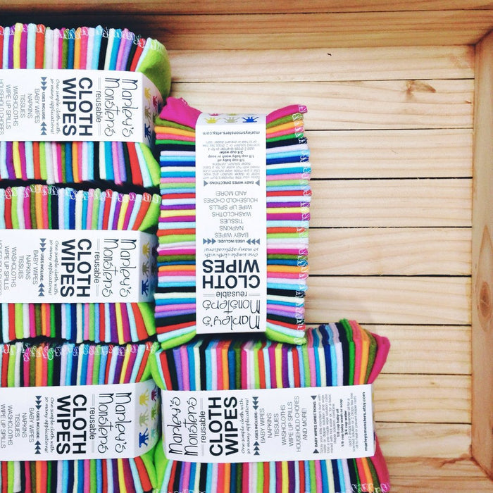 Marleys Monsters Cloth Wipes, crafted from cotton flannel with colorful stripes, are neatly stacked on a wooden surface. Each handmade wipe is packaged with a label showing the product name and details.