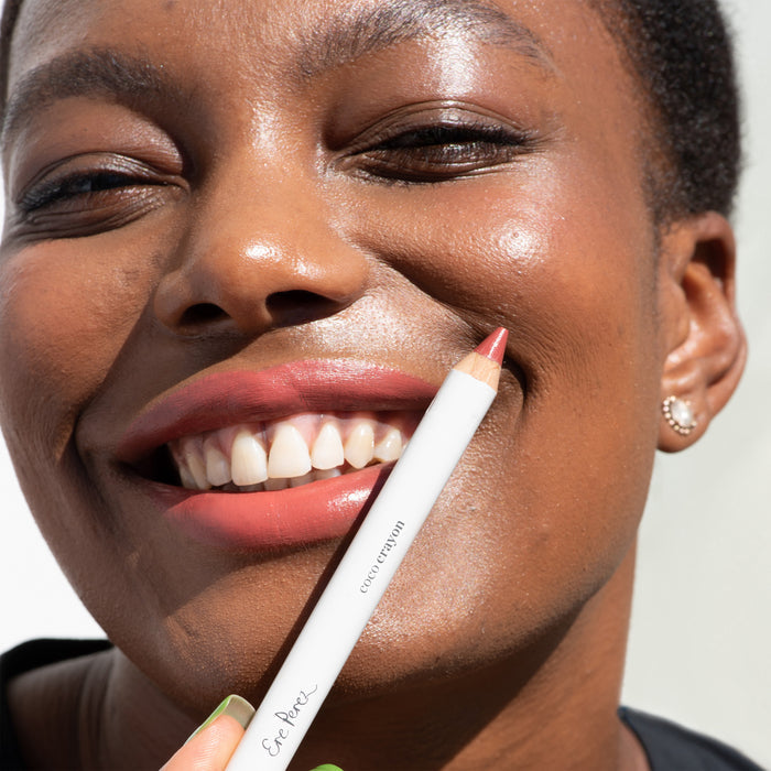 A smiling individual with short hair and small earrings applies Ere Perez Coco Crayon lip liner in coral pink, enhancing their look with its hydrating creamy formula against a soft white background. They look directly at the camera, showcasing the natural beauty crayon.