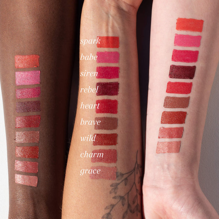 Three arms with diverse skin tones showcase Ere Perezs Coco Crayon zero-waste lipsticks in shades like Spark, Babe, and Siren. The creamy, hydrating formula includes pinks, reds, browns, and nudes—perfect for natural beauty crayon lovers.