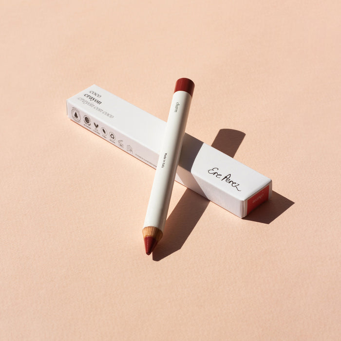 A white and red Coco Crayon from Ere Perez stands diagonally on its box against a light peach background. The brands name is visible, highlighting its hydrating creamy formula, with shadows creating a soft contrast.