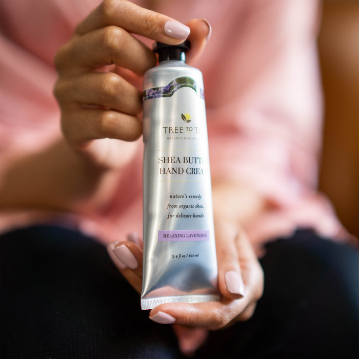 A person with light pink manicured nails holds a tube of Tree To Tubs Cocoa & Shea Butter Anti-Aging Hand Cream, creating a soothing scene in a blurred indoor setting.