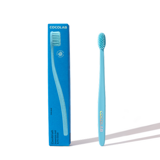 A blue Cocobrush Toothbrush by COCOLAB with soft bristles stands upright beside its matching blue box, which displays the toothbrush image and the COCOLAB brand name in white text.