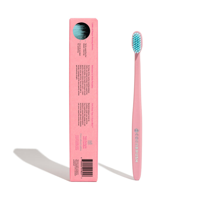 A pink Cocobrush Toothbrush by Cocofloss with a light blue bristle head stands upright beside its matching pink box. The box, featuring black text and an illustration on top, highlights the brushs signature multi-density bristles, casting a shadow on the white background.