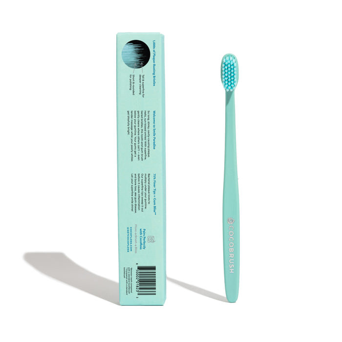 A light teal Cocobrush Toothbrush by Cocofloss stands upright beside its matching box, displaying signature brush details. Both cast shadows. The toothbrush has soft, multi-density bristles and a sleek design ideal for promoting healthy gums.