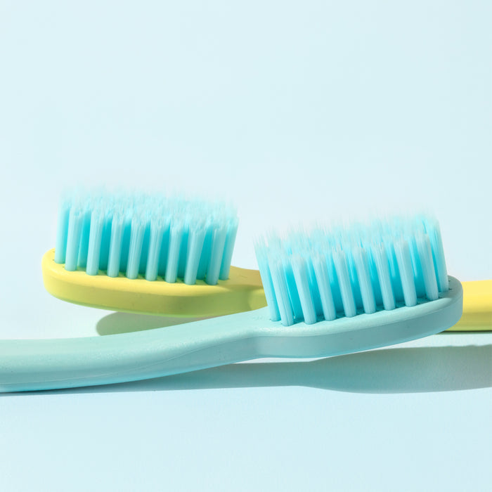 Close-up of two Cocobrush Toothbrushes by Cocofloss, featuring soft, multi-density light blue bristles for healthy gums. Set against a light blue background, one toothbrush has a turquoise handle and the other yellow.