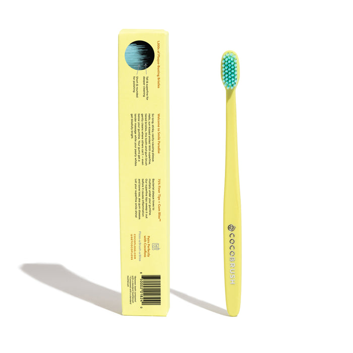 A yellow Cocobrush toothbrush with blue multi-density bristles lies beside its matching box displaying product info and the brand Cocofloss. This signature brush promotes healthy gums, set against a plain white background.