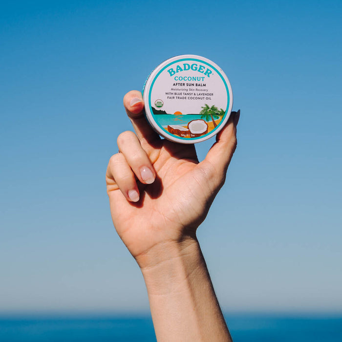 A hand raises a tin of Badger Balms Coconut After Sun Balm against a blue sky. The round tin features coconut and palm tree illustrations, emphasizing its soothing benefits from coconut oil and organic shea butter, while highlighting the products natural attributes.