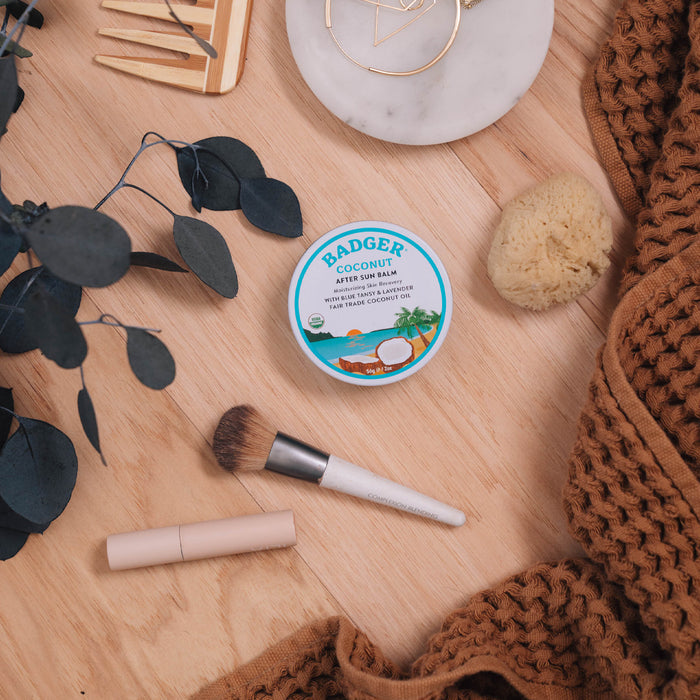 Flat lay of a wooden surface featuring Badger Balms Coconut After Sun Balm Tin with nourishing coconut oil, alongside a makeup brush, lipstick tube, textured brown fabric, eucalyptus leaves, gold earrings on a decorative plate, comb, and natural sponge.