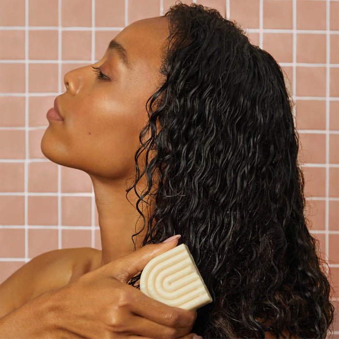 A person with wet, curly black hair uses Kitschs Coconut Oil Deep Moisturizing Conditioner Bar against a backdrop of light pink tiles.