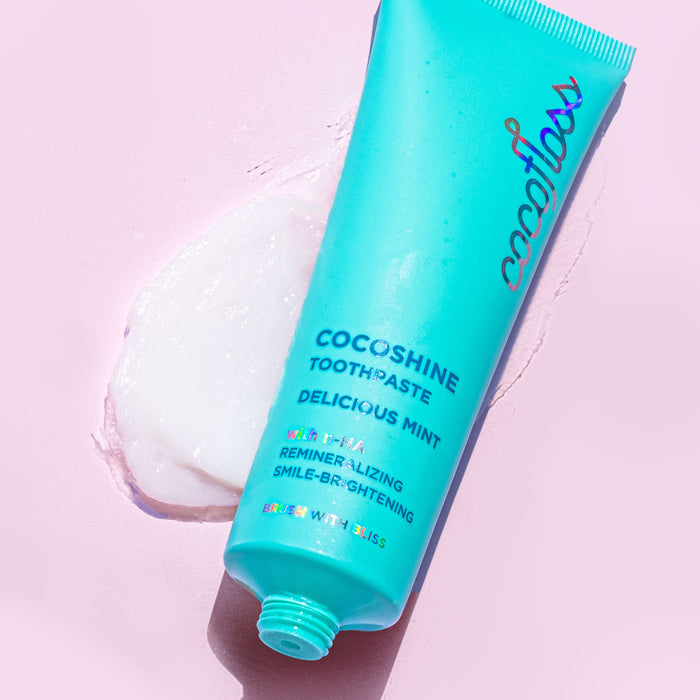 A turquoise tube labeled Cocofloss Cocoshine Whitening Toothpaste rests on a light pink surface with white paste spilling out. This toothpaste promotes enamel strengthening, remineralizing, and smile-brightening benefits with nano-hydroxyapatite.