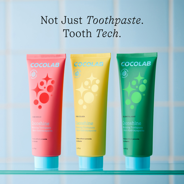 Three colorful tubes of COCOLAB Cocoshine Whitening Toothpaste (pink, yellow, and green) stand upright on a blue surface, with "Not Just Toothpaste. Tooth Tech." above them and a pale blue tiled background.