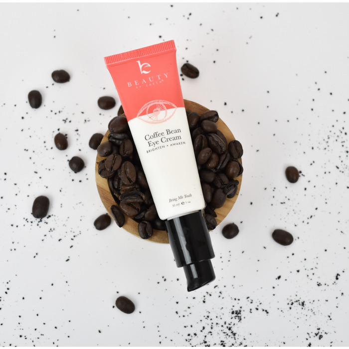 A tube of Beauty by Earths Coffee Bean Awakening Eye Cream, enriched with coffee seed extract, sits on a wooden dish amid scattered coffee beans. The under-eye cream has a red and white design and a pump dispenser, promising effective dark circle reduction.