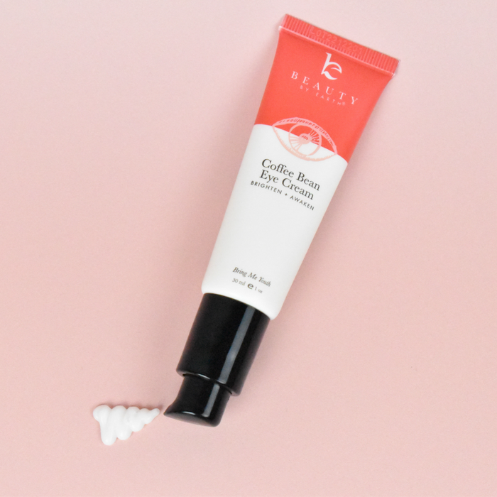On a pink surface rests the Coffee Bean Awakening Eye Cream by Beauty by Earth. The red and white tube with a black cap is enriched with coffee seed extract, and a small dollop nearby promises to reduce dark circles.