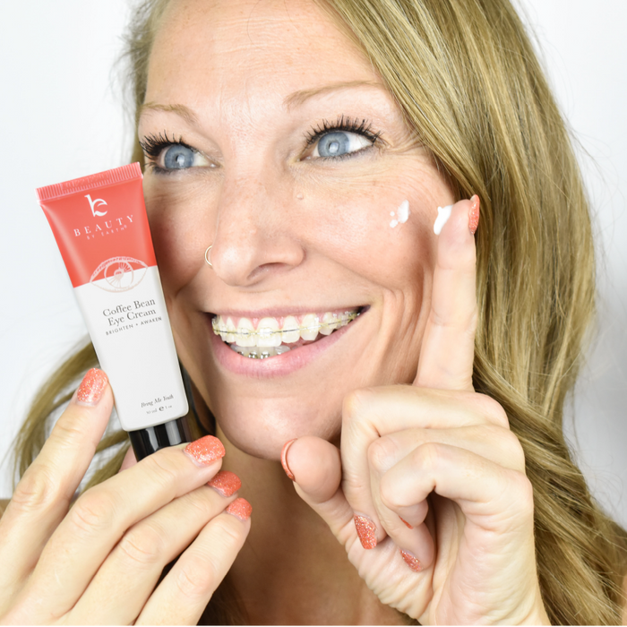 A smiling woman with braces holds a tube of Coffee Bean Awakening Eye Cream by Beauty by Earth, celebrated for its coffee seed extract that helps reduce dark circles. She applies a small amount under her bright blue eyes, while her orange-painted fingernails add a pop of color in her demonstration.