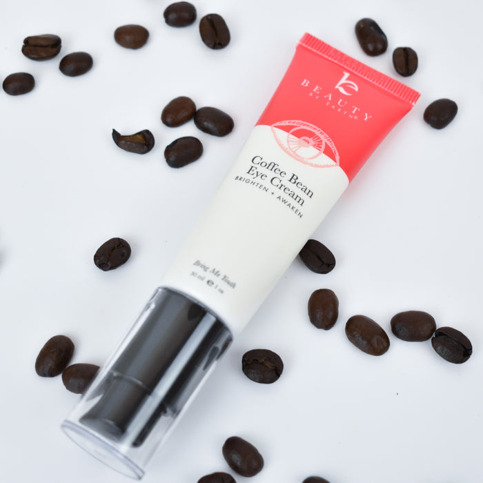 A Beauty by Earth Coffee Bean Awakening Eye Cream tube rests on a white surface, surrounded by scattered coffee beans. The pink and white tube, adorned with a round graphic and the brand name, contains coffee seed extract to reduce dark circles and rejuvenate tired eyes.