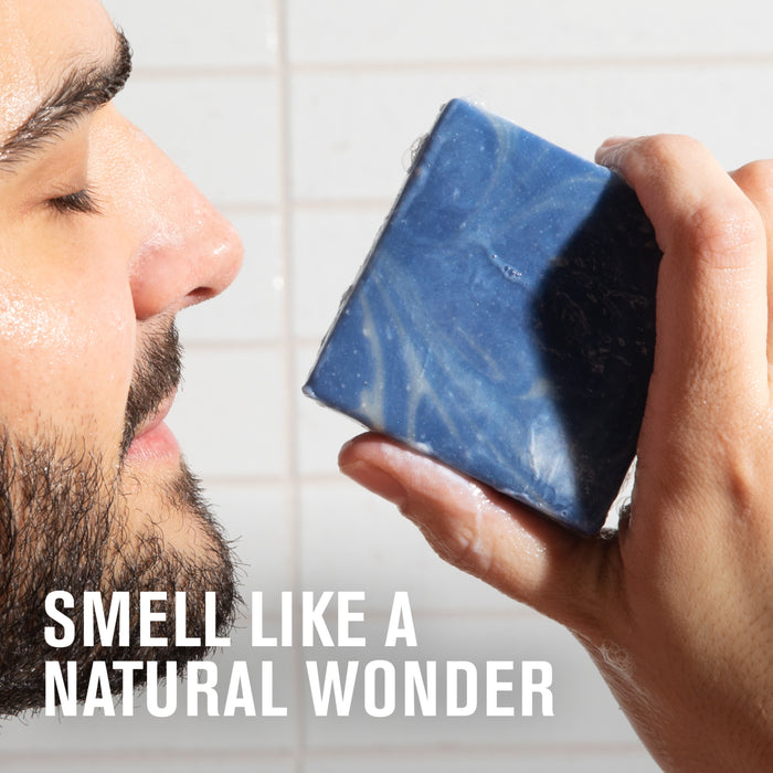 A man holds a blue Every Man Jack Cold Plunge Bar Soap close to his nose, eyes closed, savoring the scent. The text reads: SMELL LIKE A NATURAL WONDER. White tiled wall in the background.