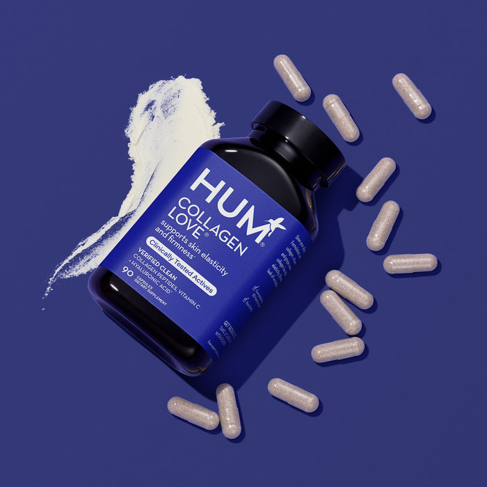 A bottle of HUM Nutritions Collagen Love rests on a blue surface, surrounded by scattered capsules. Next to it, a white powder smear is visible. The label emphasizes collagen peptides and vitamin C as key ingredients to enhance skin elasticity and reduce fine lines.