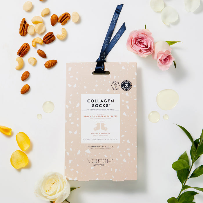A box of Collagen Socks Trio by VOESH New York, featuring a vegan formula, is surrounded by rose petals, nuts, oil droplets, and fresh flowers. With a ribbon-adorned package, it showcases moisturizing foot masks with argan oil and floral extracts.