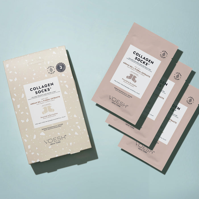 A flat lay features the VOESH New York Collagen Socks Trio packaging—a speckled beige box with bold white text—and three light pink sachets on a light blue background. The vegan formula includes foot masks enriched with Argan Oil and Floral Extracts.