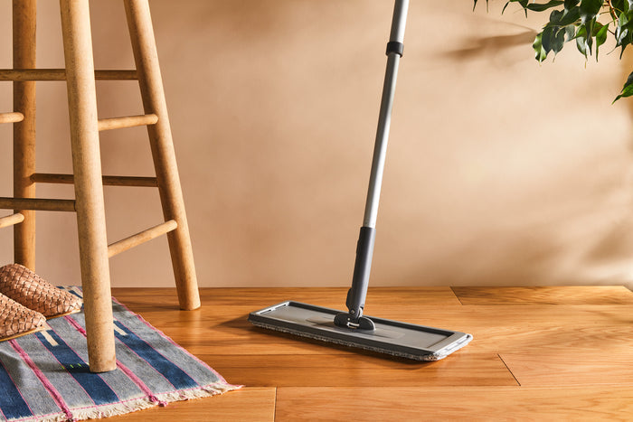 The Collapsible Universal Handle by Grove Co., a modern mop with a flat head, rests beside a wooden chair on the floor. Made from recycled materials, it perfectly complements the woven rug and green plant leaves, creating a clean and cozy atmosphere.