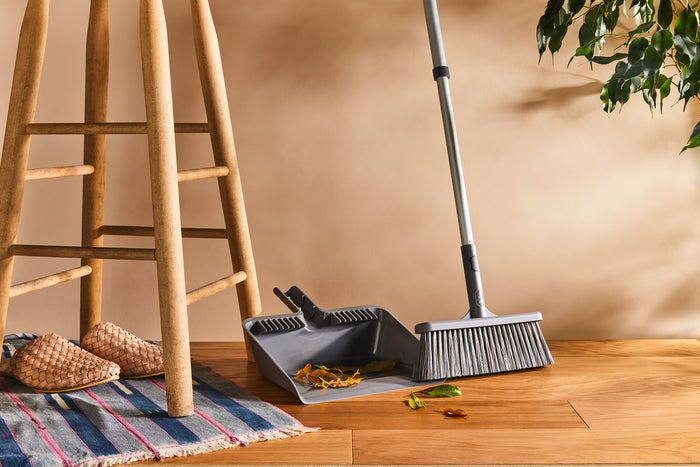 A Grove Co. Collapsible Universal Handle broom with a dustpan is on a wooden floor beside a stool with woven slippers on a striped rug. A green plant against the beige wall highlights the charm of recycled materials in harmonious living spaces.