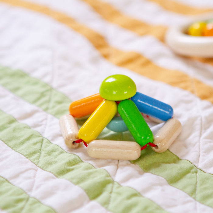 The Color Carousel Wooden Baby Rattle by HABA is showcased on a quilt with green, white, and yellow stripes. The multicolored clutching toy has interconnected wooden pieces in vivid green, blue, yellow, and orange hues arranged in a circular design.