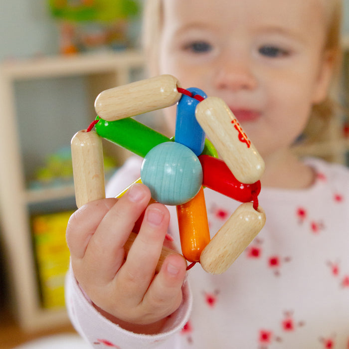 A toddler wearing a white shirt with red patterns holds the HABA Color Carousel Wooden Baby Rattle, featuring a central blue sphere and colorful rods in red, orange, yellow, and green. The blurred background shows shelves filled with books and toys.