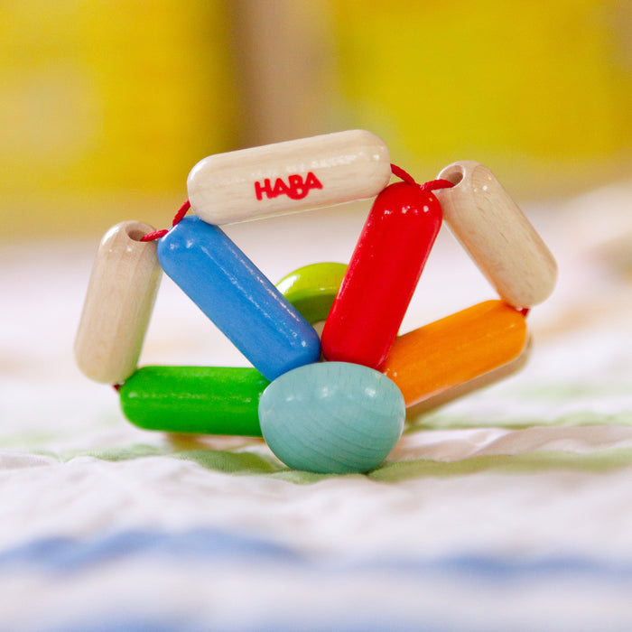The HABA Color Carousel Wooden Baby Rattle is a vibrant toy with interconnected red, blue, green, and orange rods. It sits on a soft, striped surface against a blurred yellow background.