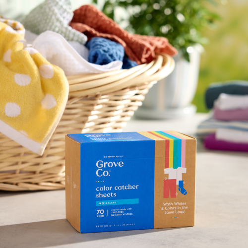 Grove Co. Color Catching Laundry Sheets sit prominently on the table, ideal for any laundry day. A wicker basket holds freshly folded laundry in vibrant yellow, blue, and white hues against a backdrop of softly blurred greenery, enhancing this tree-free solution scene.