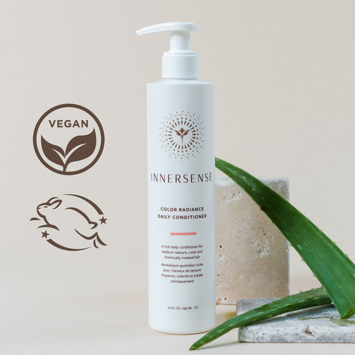 A bottle of Innersense Color Radiance Daily Conditioner next to aloe leaves and a stone slab. The bottle features vegan and cruelty-free symbols. The background is a soft beige surface.